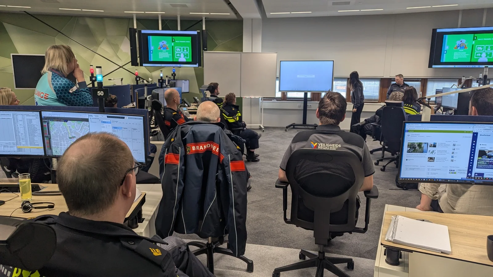 Brandweer-leerlokaal &mdash; uniformen op de voorgrond, presentatie op groot scherm, mensen luisteren mee.