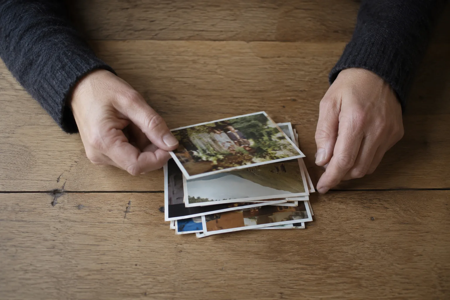 Twee handen die door een kleine stapel oude familiefoto's bladeren op een warm-houten tafel, daglicht van links.