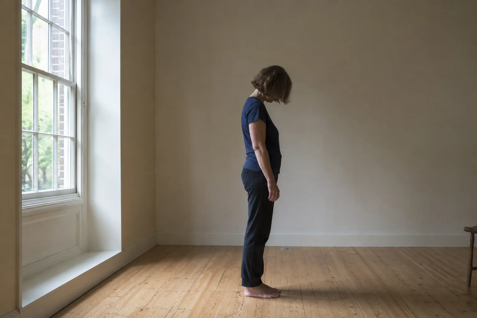 Een vrouw staat in een lichte praktijkruimte op blote voeten van zij-aanzicht, hoofd licht omlaag, daglicht door een groot raam.